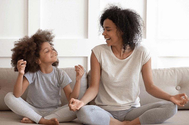 A mother and young daughter sitting on a couch holding a yoga pose.